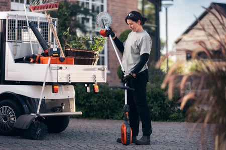 Husqvarna 525iLK kombimotor – uden redskab og batteri – En person, der holder et haveredskab ved siden af et køretøj, der er udstyret med forskellige haveprodukter.