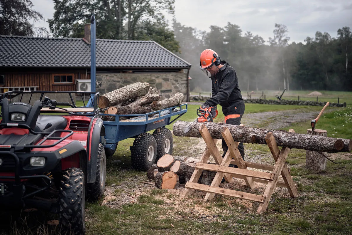 Husqvarna 435i motorsav – 35 cm, uden batteri – En person i arbejdsudstyr, der bruger en kædesav til at skære kævler på en savbuk ved siden af en quadcykel og anhænger.