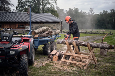Husqvarna 435i motorsav – 35 cm, uden batteri – En person i arbejdsudstyr, der bruger en kædesav til at skære kævler på en savbuk ved siden af en quadcykel og anhænger.