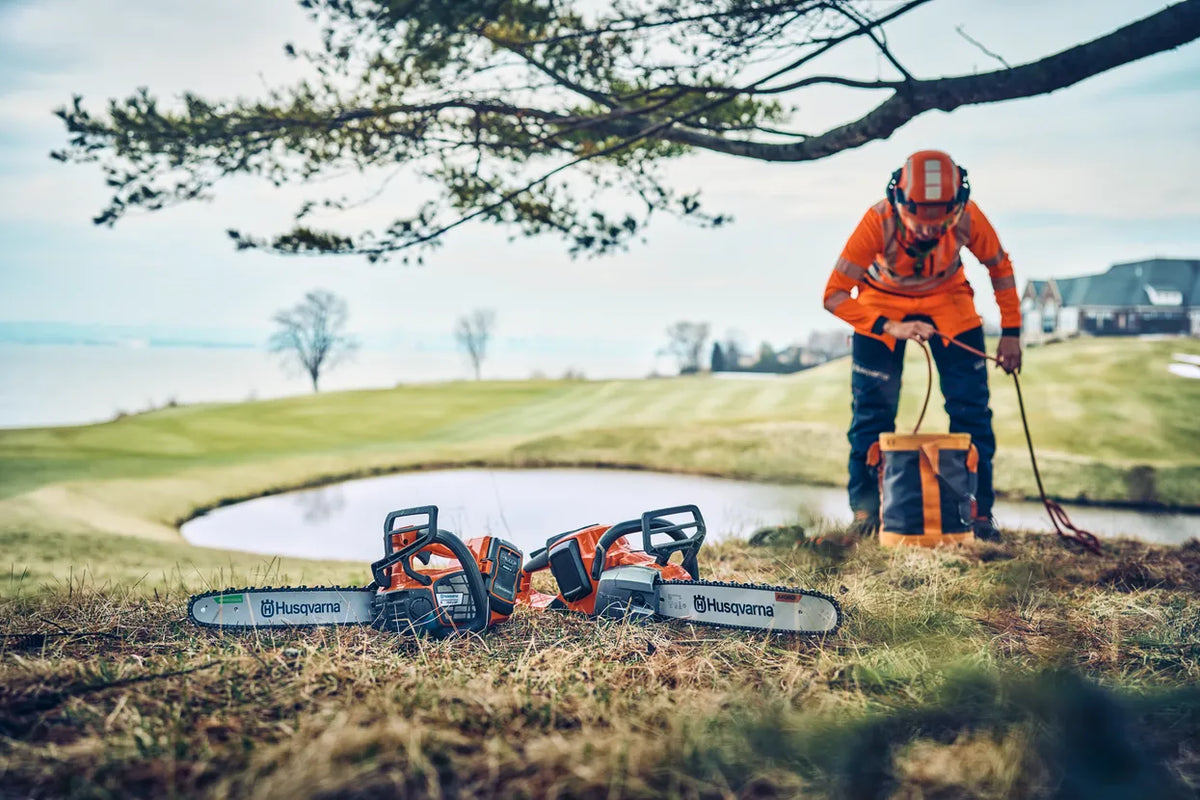 Husqvarna 542i XP G motorsav – 33 cm, uden batteri – En person i arbejdsudstyr, der opsætter udstyr nær en dam med to Husqvarna-motorsave på græsset.