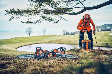 Husqvarna 542i XP G motorsav – 33 cm, uden batteri – En person i arbejdsudstyr, der opsætter udstyr nær en dam med to Husqvarna-motorsave på græsset.