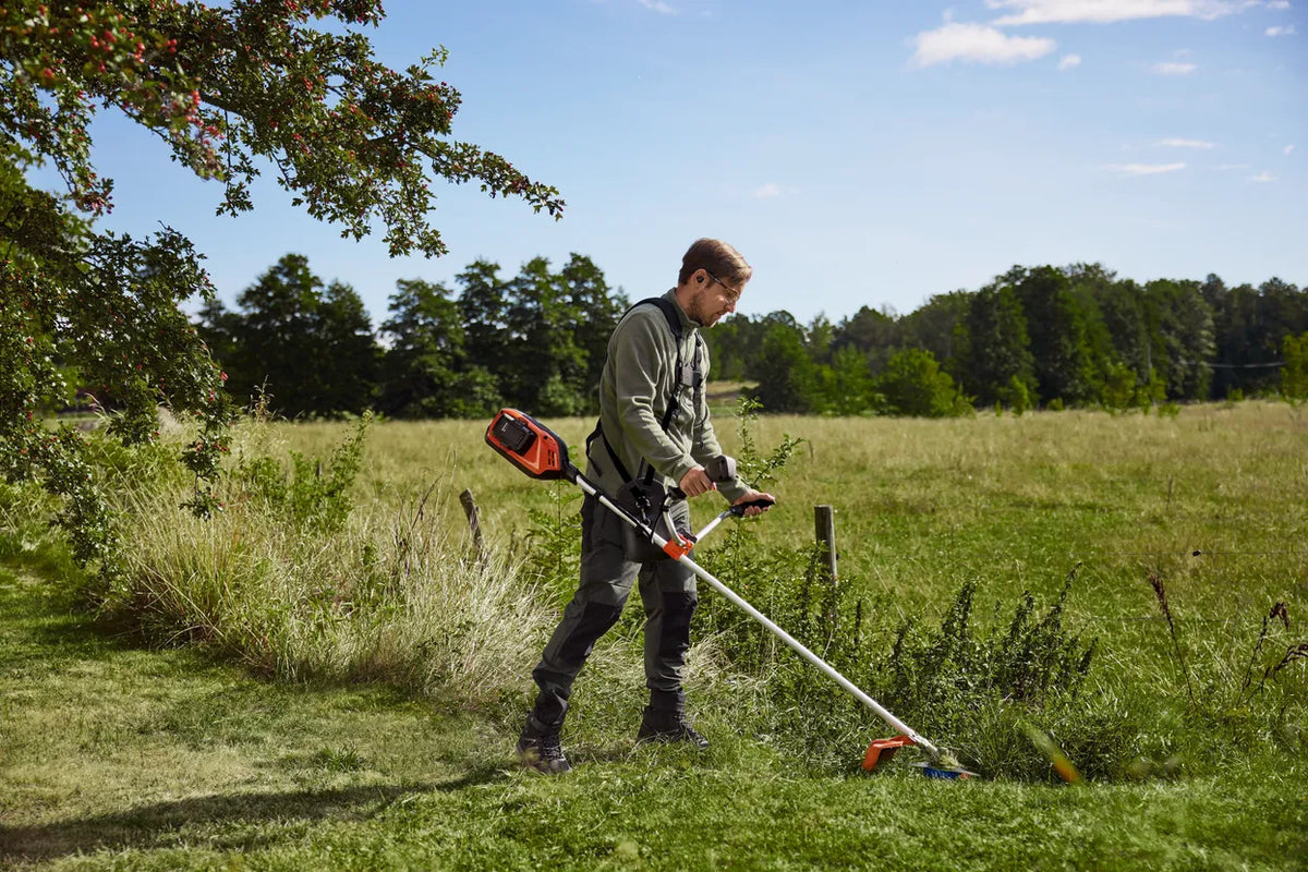 Husqvarna 325iR batteribuskrydder – uden batteri – En person, der bruger en trådtrimmer til at klippe græs langs en landlig hegnslinje.
