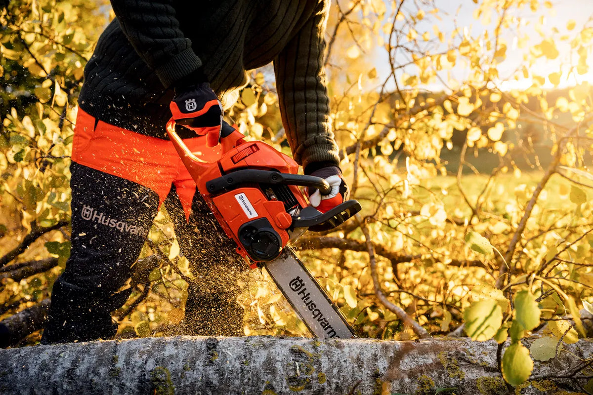 Husqvarna Classic 20A savbukser – str. 44, klasse 1 – En person, der bruger en Husqvarna-motorsav til at save en nedfalden gren i skoven.