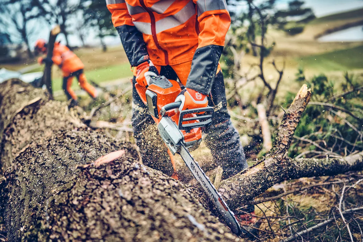 Husqvarna 542i XP G motorsav – 33 cm, uden batteri – En person, der bruger en kædesav til at save i en nedfaldet træstamme udendørs med en anden person i baggrunden.