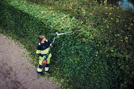 Husqvarna 525iHE3 stanghækkeklipper – uden batteri – En person, der bruger en hækkeklipper til at trimme toppen af en tæt hæk i en have.