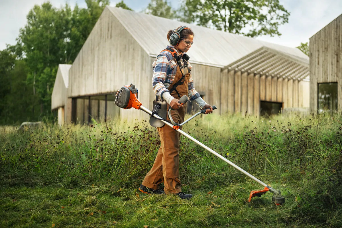 Husqvarna 129R buskrydder med trimmerhoved og klinge – En person, der bruger en buskrydder til at trimme højt græs ved siden af moderne træbygninger udendørs.
